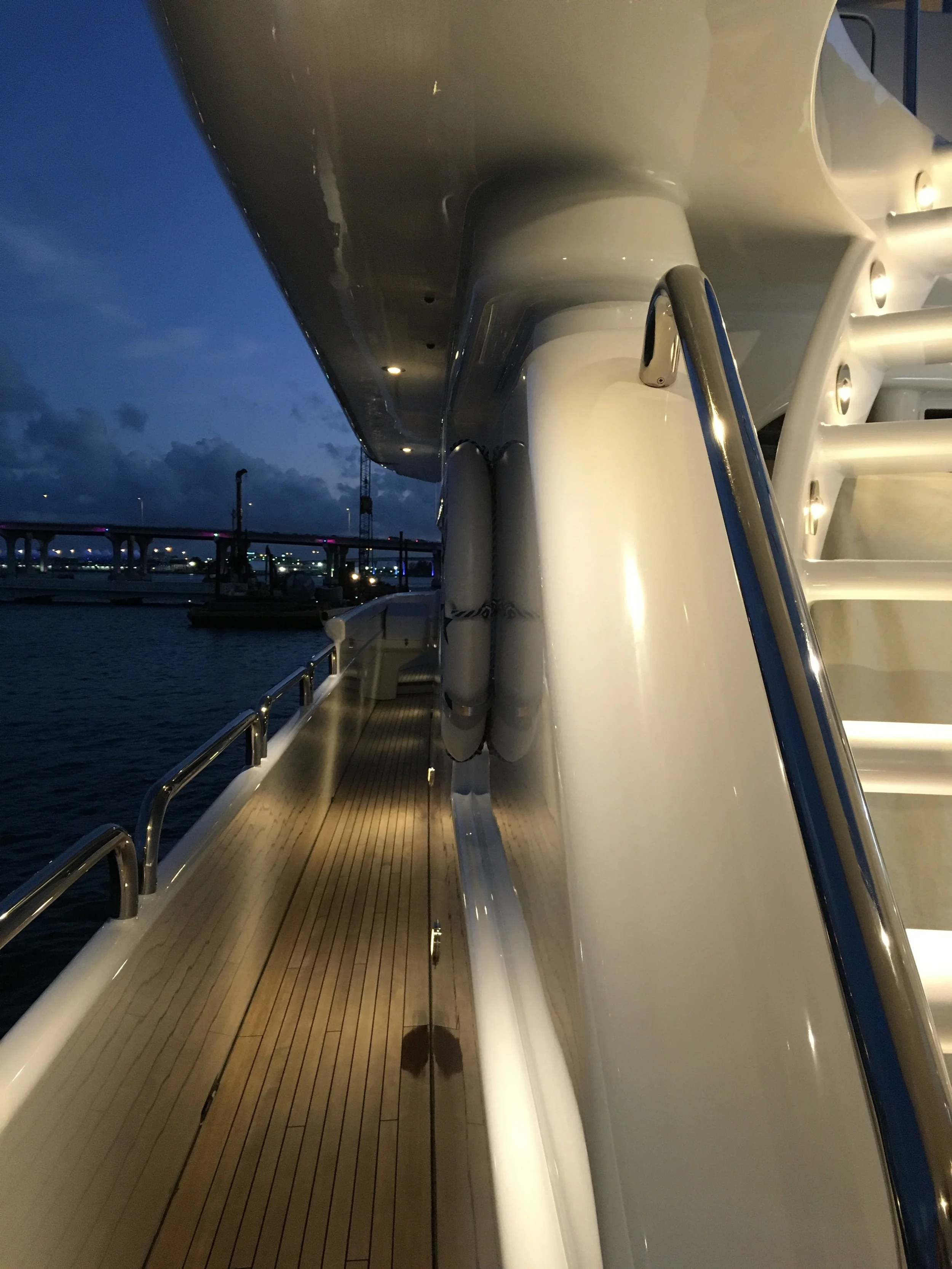 Polished teak deck of a motor yacht at dusk, chrome railings catching city lights and a bridge reflected on still water